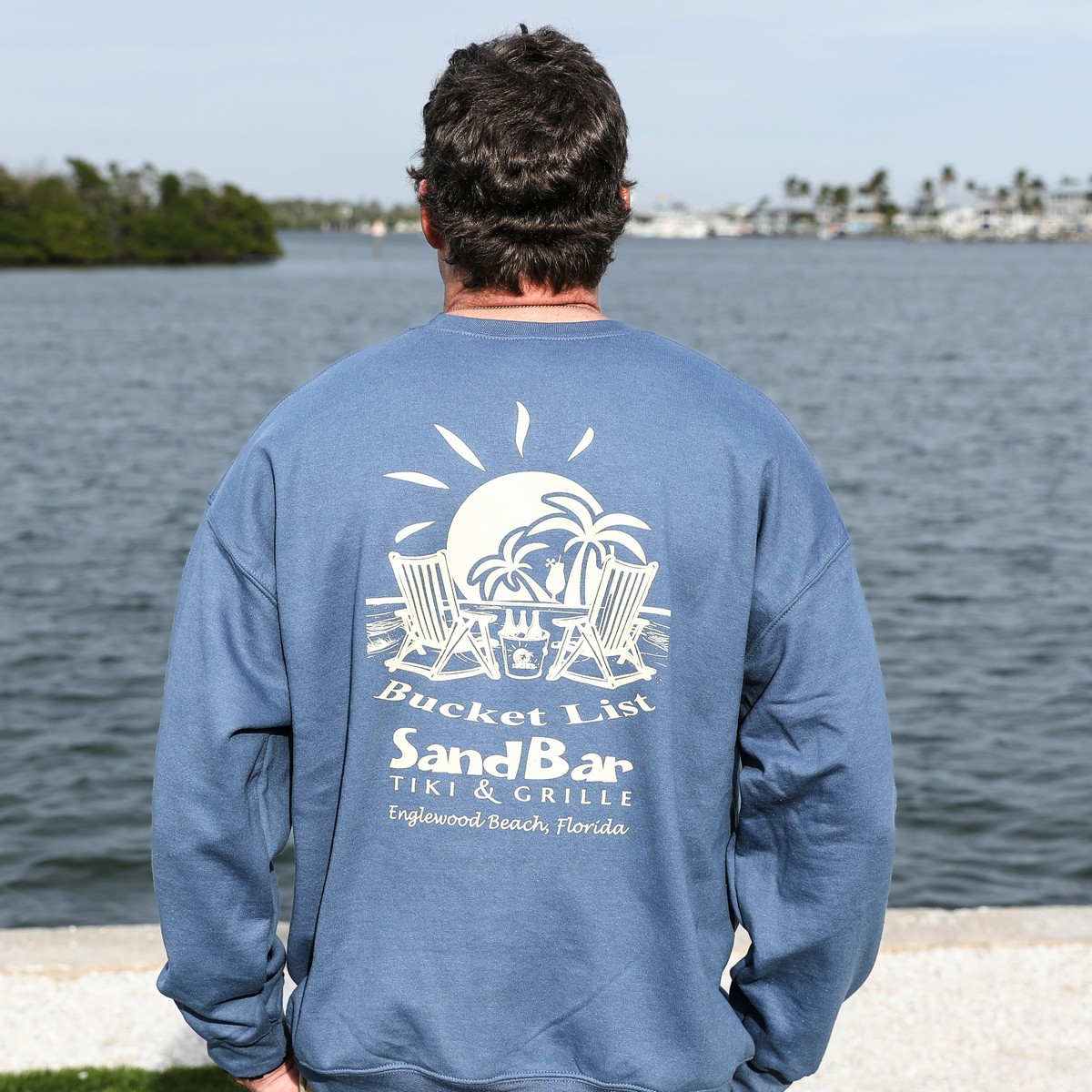 man wearing Sandbar Tiki & Grille Crewneck Sweatshirt with intracoastal waterway in the background - back view